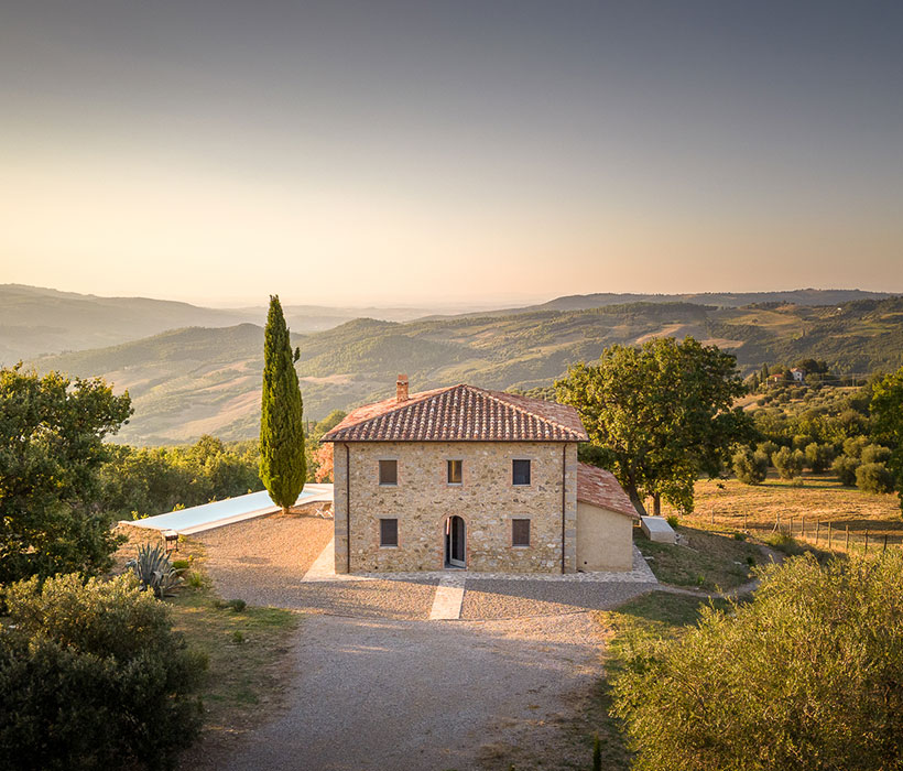 Casa colonica in pietra con tetto in tegole, circondata da colline e alberi, vista panoramica rurale.