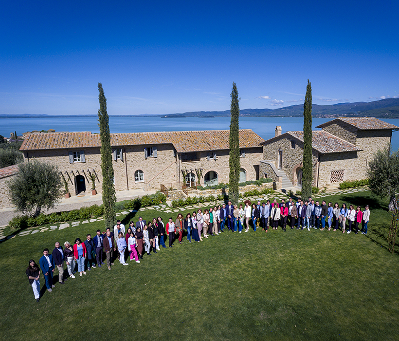 Ampia villa in pietra con tetto in tegole vicino al lago, giardino curato e gruppo di persone sul prato.