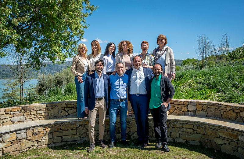 Gruppo di persone posa all'aperto su una panchina di pietra con verde e lago sullo sfondo.