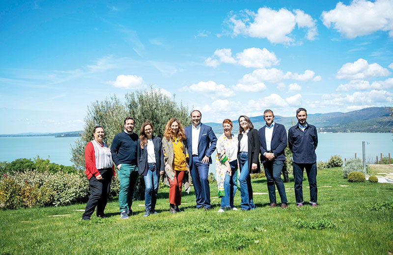 Gruppo di persone posa su un prato con lago e montagne sullo sfondo.
