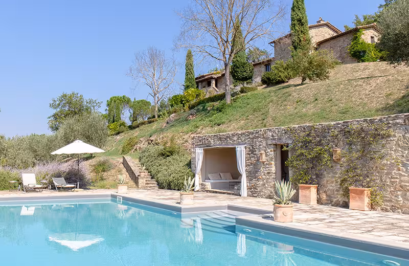Elegante villa in pietra con piscina e giardino su una collina sotto un cielo azzurro.