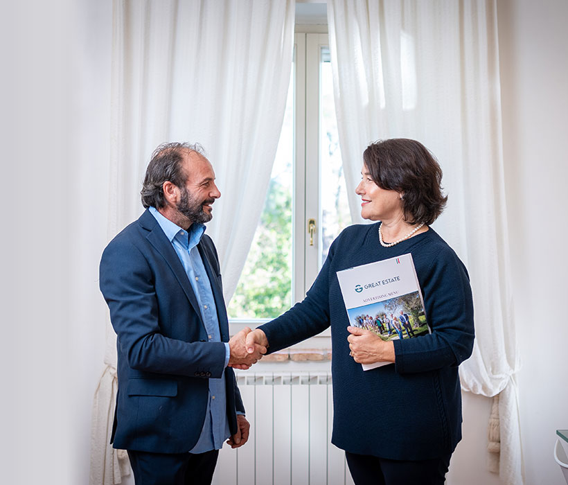 Due persone si stringono la mano in un ufficio luminoso con documenti Great Estate.