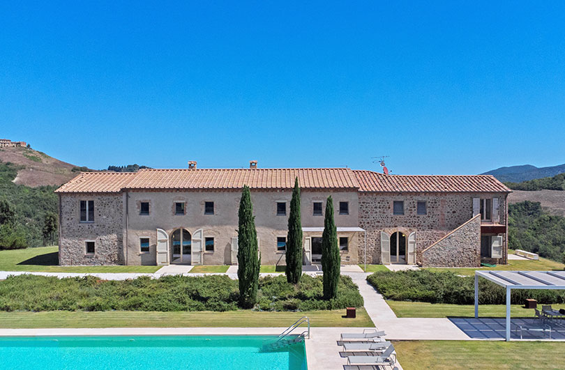 Ampia villa rustica con facciata in pietra, piscina e cipressi in campagna panoramica.