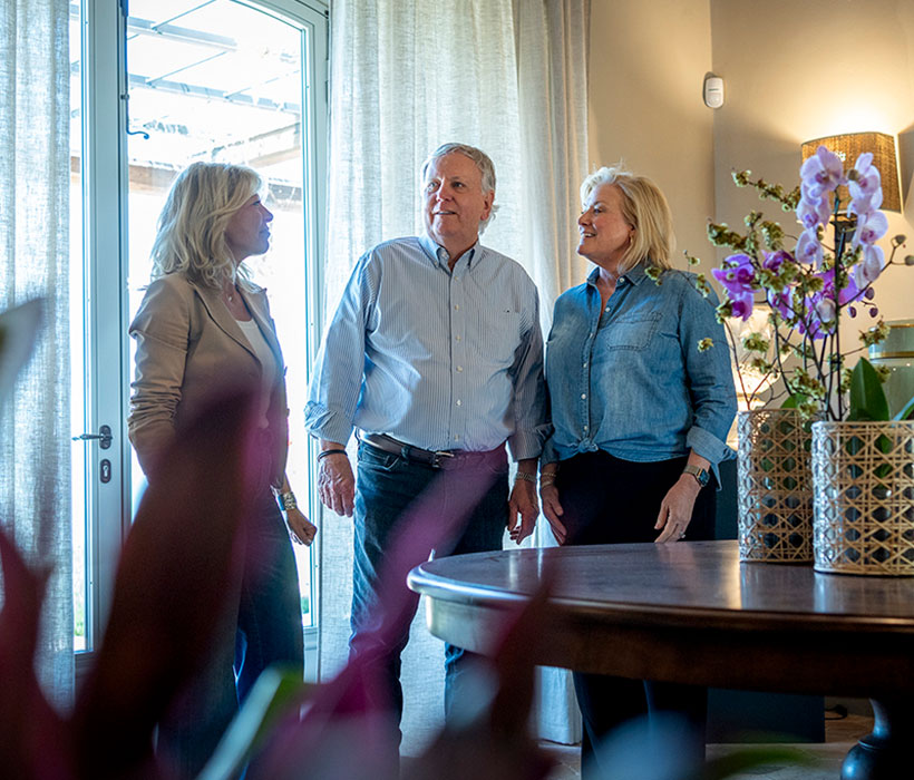 Tre persone che parlano in un luminoso soggiorno con grande finestra e fiori decorativi.